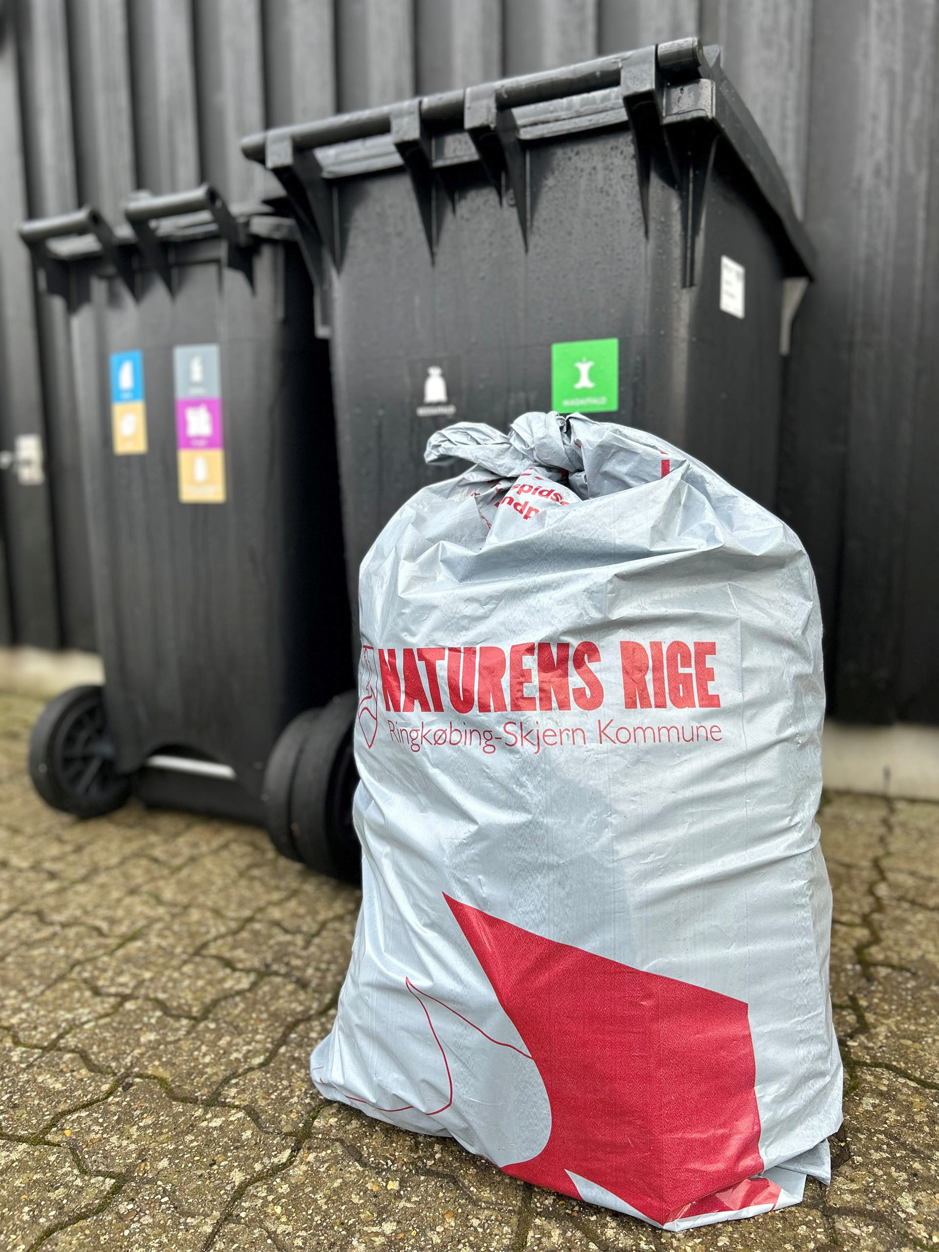 Foto af en fyldt sæk med ekstra-restaffald. Det er en officiel restaffaldssæk, som er grå med rødt Ringkøbing-Skjern Kommune-tryk. Sækken er placeret ved en mad- og restaffaldsbeholder, så skraldefolkene kan tage den med ved tømning af mad- og restaffald.